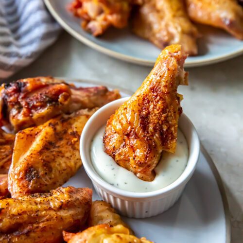 A plate of cooked chicken wings, home-cooked to perfection, with one wing dipped in a small bowl of sauce. Another plate of chicken wings is in the background.