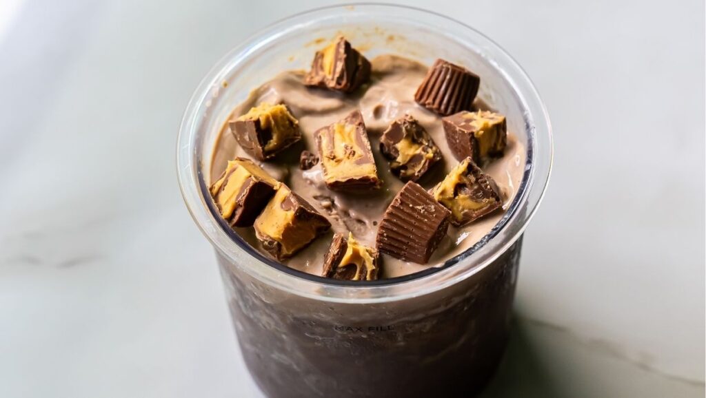 A glass filled with a rich chocolate beverage, topped with pieces of peanut butter cups, transformed into a smooth blend using the Ninja Creami for a delightful dairy-free treat.