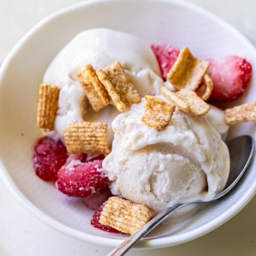 A bowl of refreshing protein ice cream topped with cereal flakes and fresh strawberries, with a spoon placed on the side.