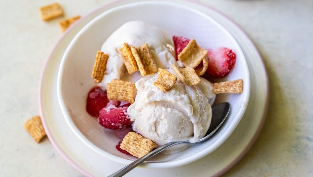 A bowl of protein ice cream topped with strawberries and cereal pieces, with a spoon on the side.