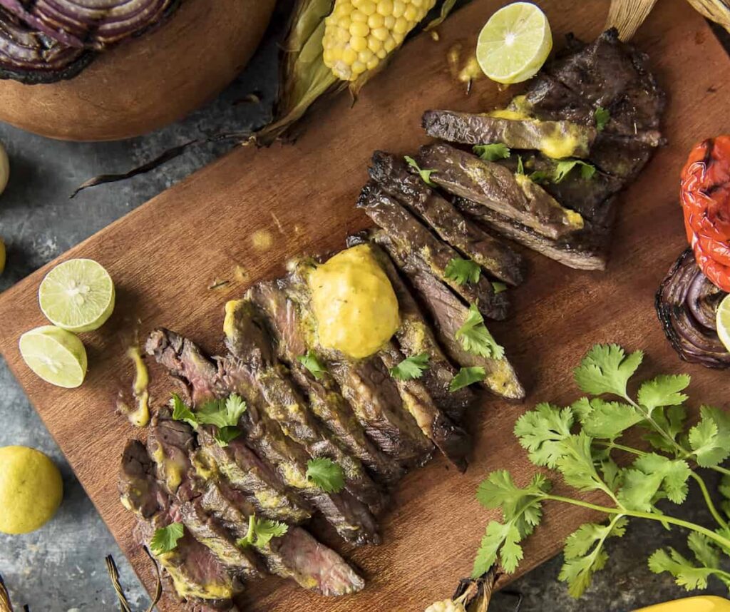Sliced grilled steak with a yellow sauce on a wooden board, garnished with cilantro, surrounded by lime halves, corn, and vegetables. Discover this dish and more in our 28 steak recipes collection.