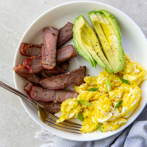 A plate showcasing a tasty steak breakfast: sliced leftover steak, scrambled eggs topped with herbs, and fresh avocado slices. A fork rests on the plate, while a gray napkin peeks from underneath.