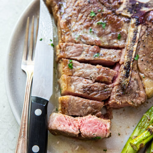 A plate with a partially sliced Porterhouse steak and cooked asparagus. A fork holds one slice of the perfectly reverse-seared steak. An orange napkin is partially visible on the lower right side.