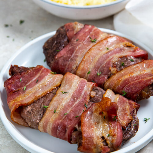 Two delicious air-fryer bacon-wrapped steaks on a white plate are garnished with herbs. A bowl of mashed potatoes is in the background.