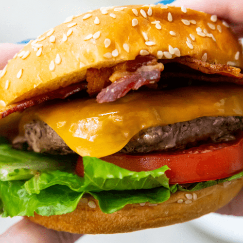 A must-have close-up of a cheeseburger with bacon, lettuce, and tomato in a sesame seed bun, being held by two hands—a perfect showcase for your Blackstone Griddle accessories.
