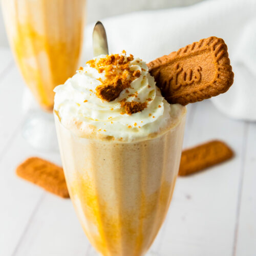 Two Biscoff milkshakes topped with whipped cream, crumbled cookies, and a whole cookie in each glass, placed on a white surface with additional cookies in the background.