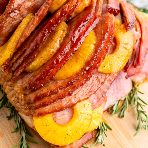 Sliced glazed ham topped with pineapple rings, a hint of brown sugar, and sprigs of rosemary rests elegantly on a wooden board.