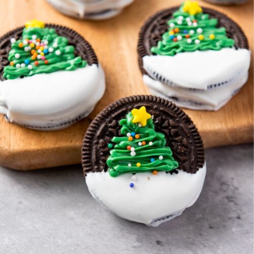 Five Oreos partially dipped in white icing, each adorned with a green icing Christmas tree and a yellow star, are placed artfully on a wooden board.