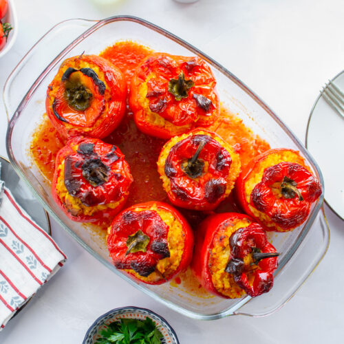 A glass dish holds six baked red bell peppers stuffed with rice, the star of this delightful stuffed pepper recipe. Nearby are plates, cutlery, napkins, and a bowl of fresh tomatoes.