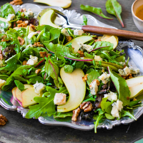 A festive harvest salad on a silver platter featuring mixed greens, pear slices, walnuts, and blue cheese. A bowl of spicy citrus vinaigrette and wooden utensils are nearby on a wooden table.