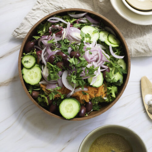 A wooden bowl brimming with a garden salad features sliced cucumbers, red onions, olives, parsley, and shredded carrots, all beautifully placed on a fabric napkin.