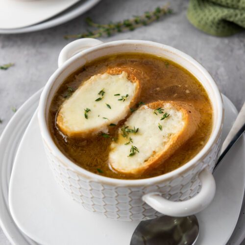 A steaming bowl of iconic French onion soup, completed with two slices of bread crowned with melted cheese and garnished with fresh herbs. A spoon elegantly rests on the white plate beneath.