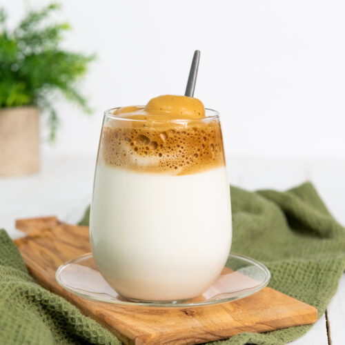 A glass of creamy Dalgona coffee with a rich foam topping, elegantly set on a wooden board with a green cloth underneath.