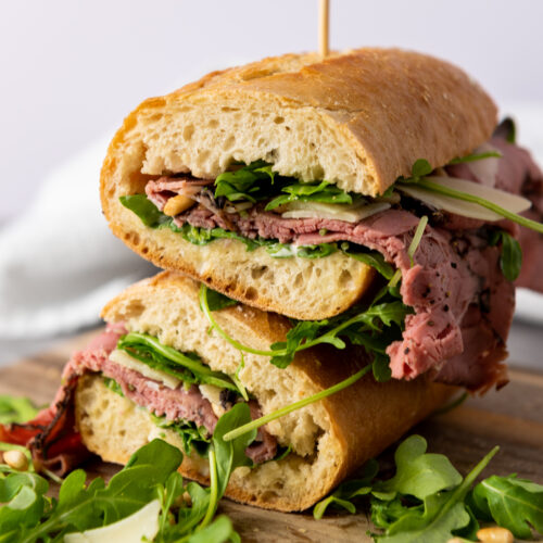 A stacked roast beef sandwich on a wooden board with arugula, cheese, and pine nuts is elevated with a hint of truffle-mayo for an extra layer of flavor.