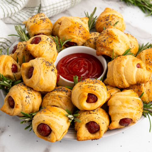 A plate of Mini Piggies in a Blanket, each sausage nestled in crescent rolls and sprinkled with poppy seeds, arranged around a bowl of ketchup. Garnished with rosemary sprigs for a touch of elegance.