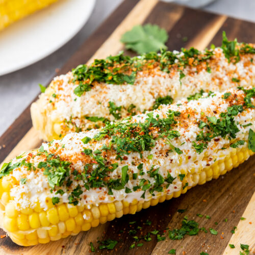 Two ears of Mexican Elotes topped with white sauce, chili powder, and chopped cilantro rest on a wooden board. A small plate with street corn and a bowl are in the background.