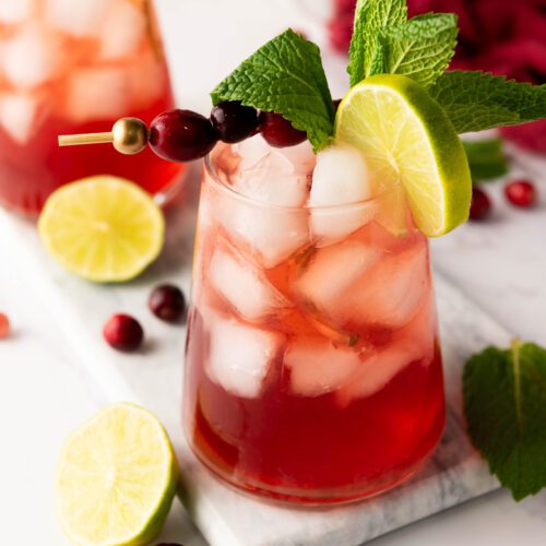 Two glasses of refreshing cranberry mocktail sit on a marble tray, their vibrant allure heightened by ice, lime slices, and mint leaves. Garnished with cranberries on skewers and surrounded by lime halves and scattered cranberries, this scene captures a perfect blend of flavors.