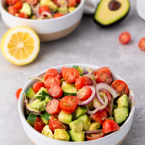 A vibrant bowl of avocado salad brimming with cherry tomatoes, cucumber, and red onion slices. Nearby, a fresh lemon and more creamy avocado rest on a sleek gray surface.