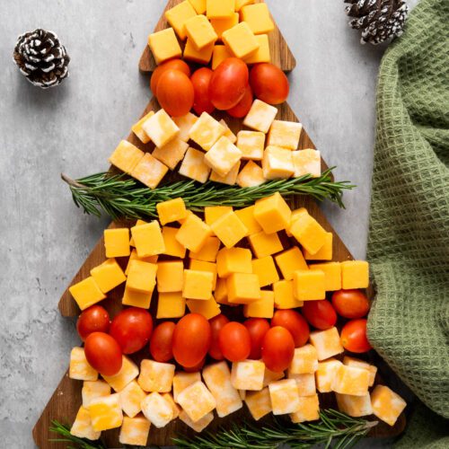 Cheese cubes and cherry tomatoes are artfully crafted into a festive Christmas tree on a wooden board, garnished with sprigs of rosemary. This delightful cheese platter is complemented by a green towel and pine cones, adding to the holiday charm.