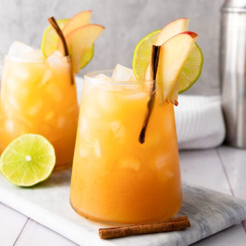 Two glasses of a light orange mocktail with ice are elegantly garnished with lime slices, apple slices, and cinnamon sticks, resting on a marble board. The subtle hint of ginger adds a delightful twist to this refreshing drink.