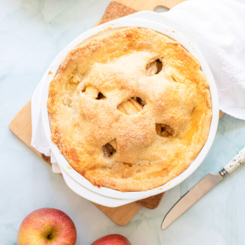 A freshly baked Honeycrisp apple pie in a white dish on a wooden board, surrounded by two whole apples, a knife, and a white napkin on a light-colored surface.