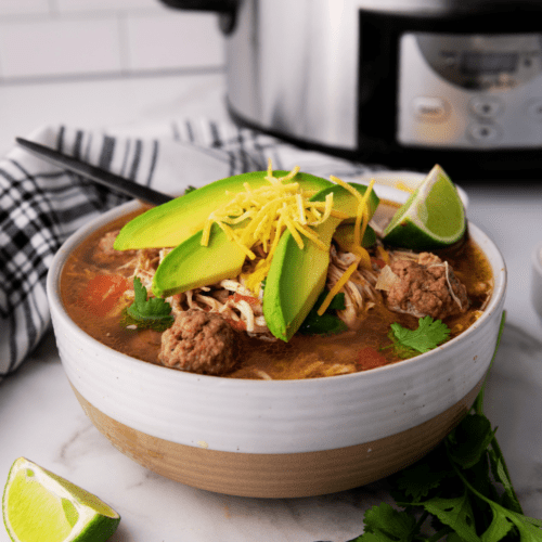 A bowl of taco soup brimming with meatballs, avocado slices, shredded cheese, and cilantro sits next to lime wedges. A trusty crockpot rests in the background, promising warm and hearty satisfaction.