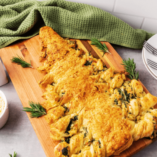 A tree-shaped puff pastry with a golden-brown crust, garnished with rosemary and spinach dip, sits on a wooden board. Plates and a green cloth are in the background, evoking the warmth of a festive Christmas Tree.