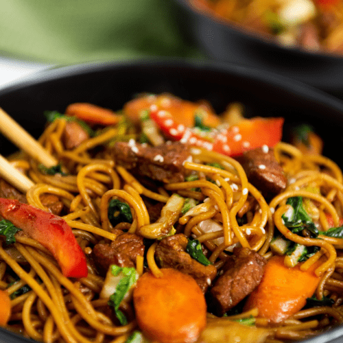 Close-up of a bowl of stir-fried lo mein with vegetables and pork, garnished with sesame seeds. Chopsticks rest on the edge of the black bowl.