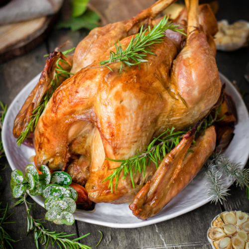 Roasted turkey, beautifully garnished with rosemary, sits on a white plate surrounded by fresh herbs and garlic on a rustic wooden table.
