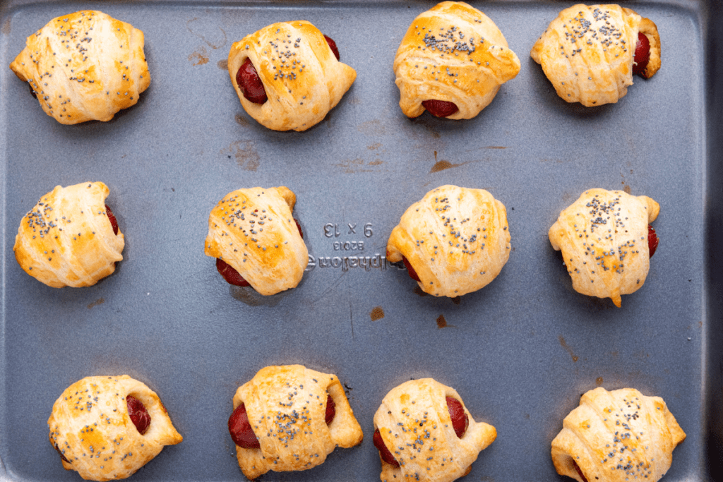 Twelve Mini Piggies in a Blanket rest on a baking tray, each topped with a sprinkle of poppy seeds.