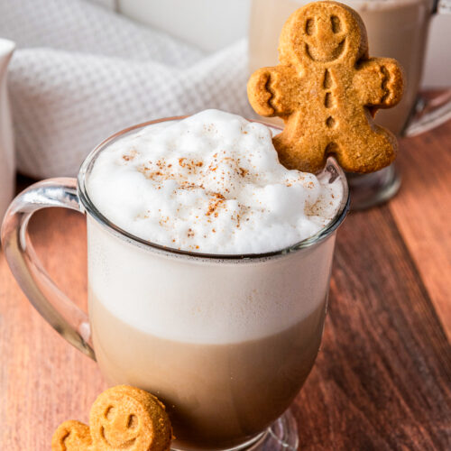 Two mugs of frothy latte topped with foam and sprinkled with cinnamon. Each mug has a gingerbread man cookie on the rim, while another cookie stands on the wooden surface nearby.