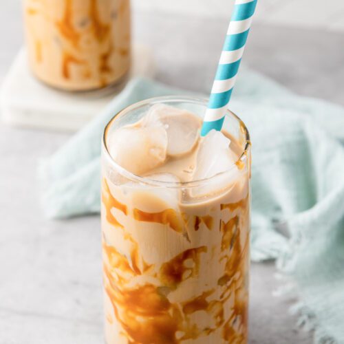 A refreshing iced caramel coffee in a glass with a blue striped straw sits on a gray surface, complemented by a light blue cloth beside it. In the background, another glass hints at the perfect coffee recipe waiting to be shared.