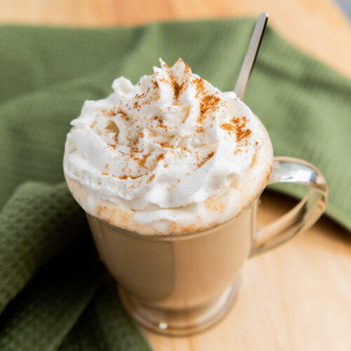 A mug of pumpkin spice latte topped with whipped cream and a sprinkle of cinnamon sits on a wooden table with a green towel.