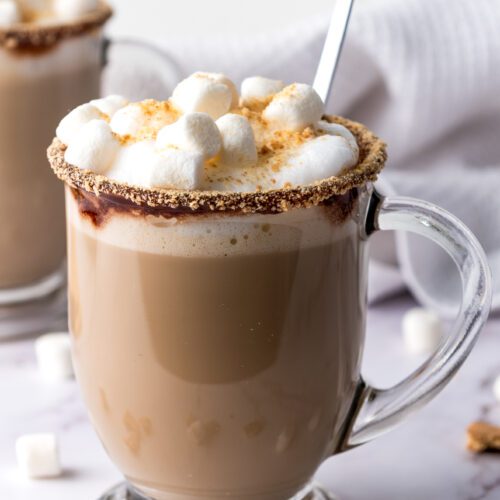A clear glass mug filled with a creamy s’mores latte, topped with marshmallows and crumbled biscuit on the rim. A spoon rests inside. Another mug and scattered marshmallows are in the background.