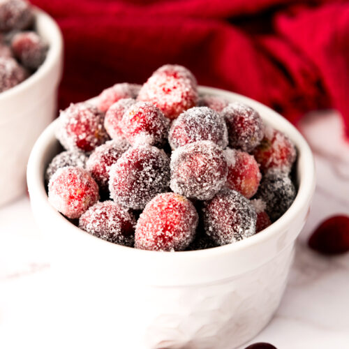 Two white bowls brimming with sugared cranberries rest on a marble surface, surrounded by a few loose berries. A red cloth serves as the backdrop.