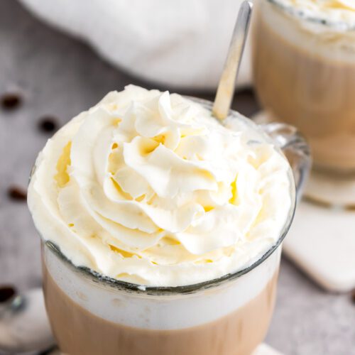 Two mugs of vanilla latte topped with whipped cream sit elegantly on coasters. A spoon is placed in one mug.