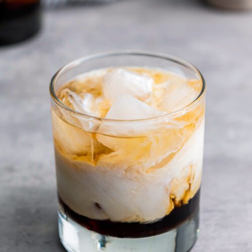 A glass of iced coffee, reminiscent of a classic White Russian, with layers of milk and coffee swirling together on a gray countertop.