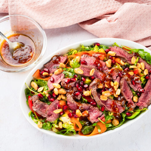 A winter salad features sliced steak, spinach, peanuts, pomegranate seeds, and tomatoes artfully arranged in a white bowl. Nearby, a small bowl of dressing waits with a spoon for the perfect finishing touch.