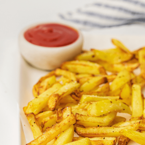 A plate of golden, crispy Air Fryer fries with a small bowl of red ketchup on the side.