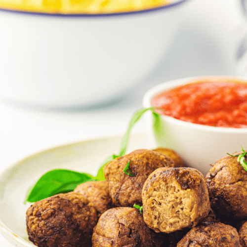 A plate of juicy air fryer meatballs garnished with fresh herbs, accompanied by a bowl of rich tomato sauce; pasta is visible in the background.