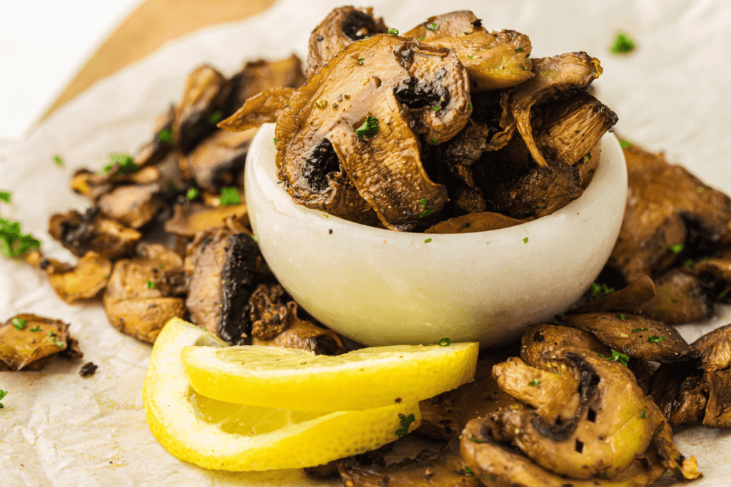 A small bowl filled with air fryer garlic mushrooms, garnished with parsley, alongside two lemon slices on a parchment-lined surface.