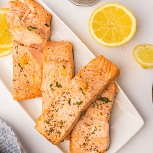 Air-fried salmon fillets on a white plate, garnished with lemon slices and herbs, with a fork and a blue napkin nearby.