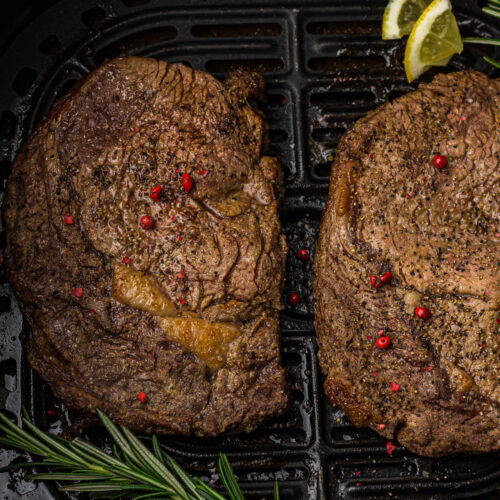 Steak cooked with peppercorns in an air fryer basket, garnished with rosemary sprigs and lemon wedges.