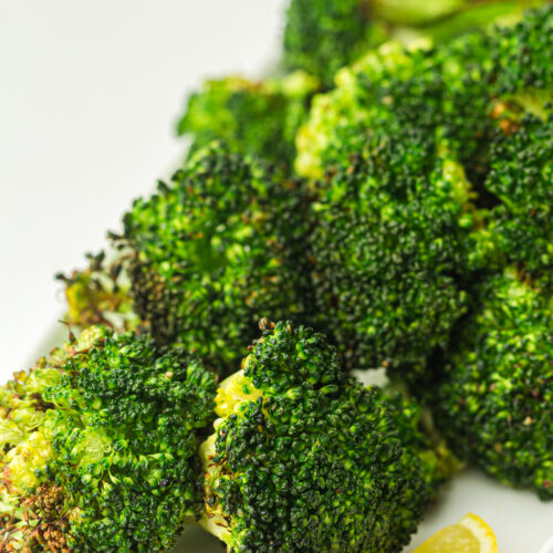 Close-up of perfectly roasted broccoli florets, effortlessly crisped in an air fryer, resting on a white plate with fresh lemon wedges.