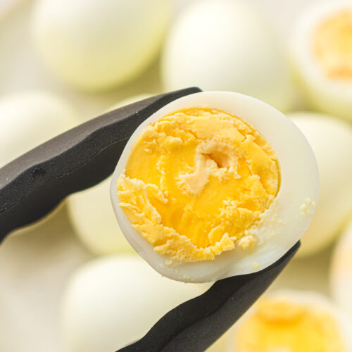 Tongs gently hold a halved hard-boiled egg with a vibrant yolk, set against a blurred backdrop of whole eggs on a plate. Perfectly cooked without the fuss, these air fryer eggs showcase simplicity and flavor at its best.