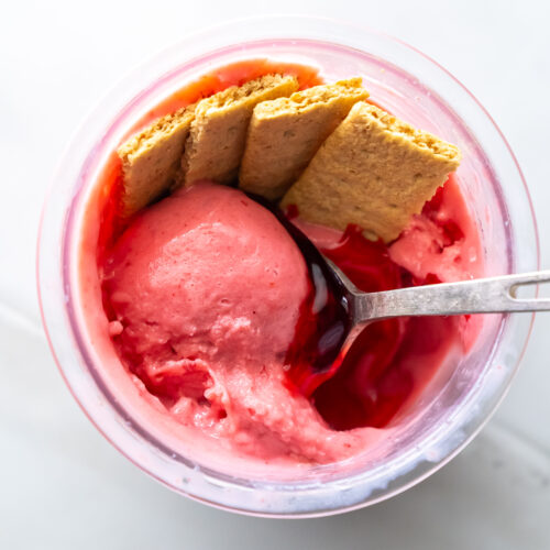 A cup of pink ice cream with a spoon, topped with a cherry sauce and three graham cracker pieces.