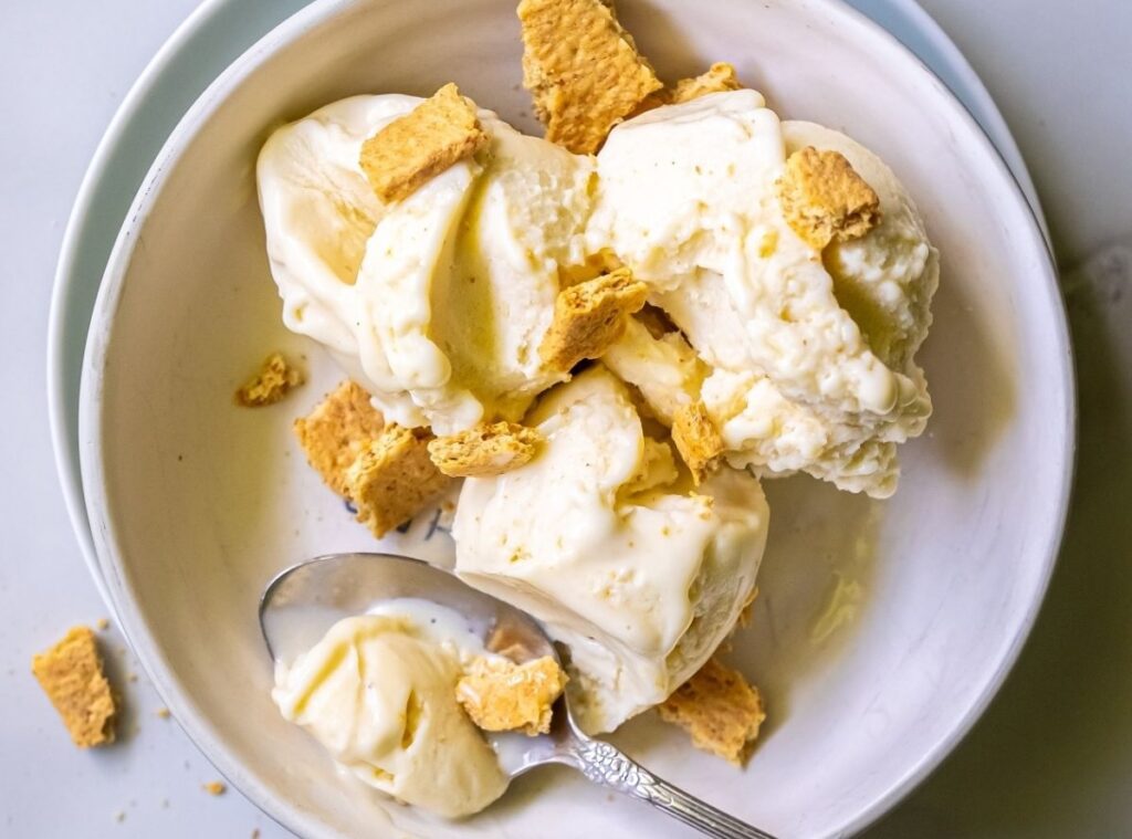 A bowl of protein-packed vanilla ice cream with scattered pieces of graham crackers, partially melted, with a spoon resting inside.