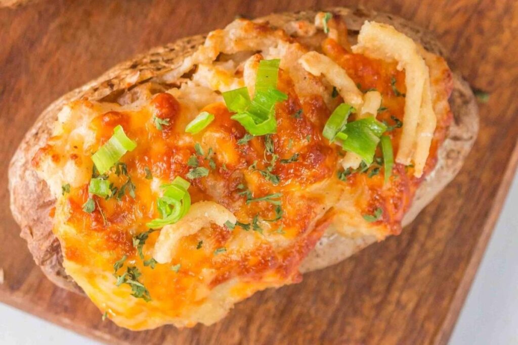 A loaded baked potato topped with melted cheese, chopped green onions, crispy onions, and herbs on a wooden surface.