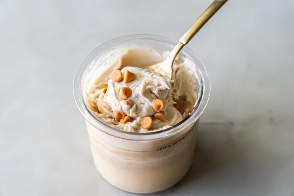 A container of creamy high-protein ice cream topped with peanut butter chips, with a gold spoon dipped into it, perfect for your cheat day indulgence.