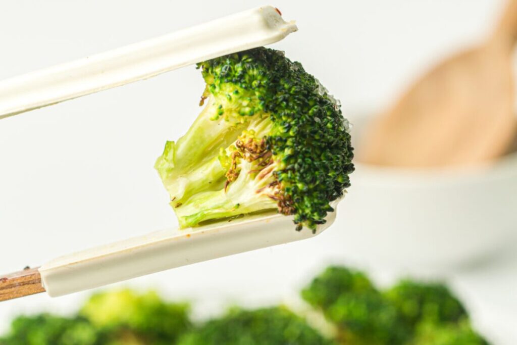 Close-up of chopsticks holding a piece of cooked broccoli, with a blurred bowl in the background.